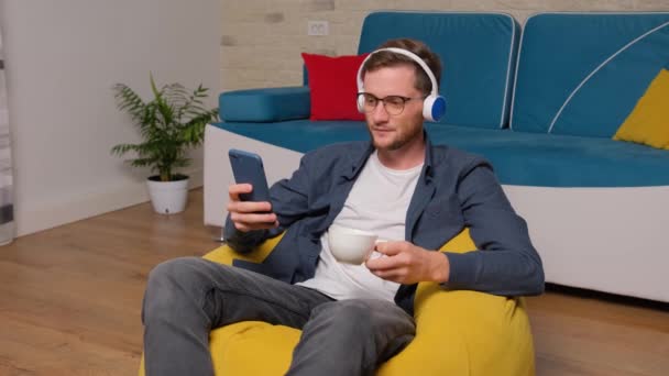 Mladý muž poslouchá hudbu na sluchátkách a popíjí šálek kávy. — Stock video