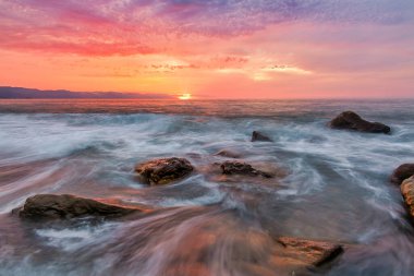 Güneş Horizon 'da batarken kayaların etrafında akan suyla rengarenk bir okyanus günbatımı