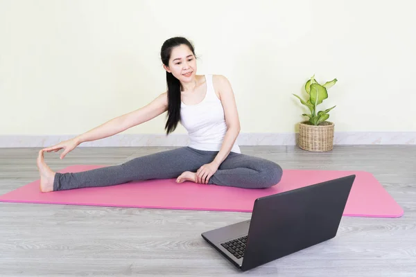 Young beautiful asian woman doing training fitness workout sport exercise feel no stress free, using laptop while sitting on floor watches a yoga tutorial at screen at home on self