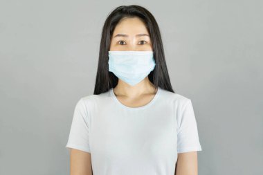 Tıbbi yüz maskesi ve beyaz tişört giyen genç Asyalı kadın gri arka planda, sağlık hizmetlerinde izole edilmiş.