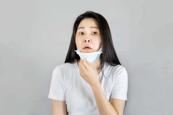 若いアジアの女性は 医療面のマスクと白いTシャツを着て彼女の手が不快な呼吸をマスクを引く 灰色の背景に隔離された不快な感じ 健康管理の概念 — ストック写真