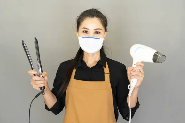 Hair salon concept,barber in medical face mask in brown apron having Hair dryer and Hair straightener in hands looking at camera, reopening of the place after the quarantine on gray background