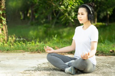 Genç güzel Asyalı kadın yoga yapıyor ve dışarıda nilüfer çiçeği pozunda meditasyon yapıyor ve kulaklıkla müzik dinliyor. Dışarıda sağlıklı bir yaşam tarzı. Yoga ve rahatlama konsepti.