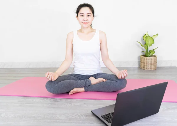 Young beautiful asian woman doing training fitness workout sport exercise feel no stress free, using laptop while sitting on floor watches a yoga tutorial at screen at home on self