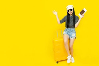 Günlük kıyafetleri ve güneş gözlükleriyle seyahat etmek için pasaportu olan genç Asyalı bir turist sarı arka planda izole edilmiş bir bagajda oturuyor.