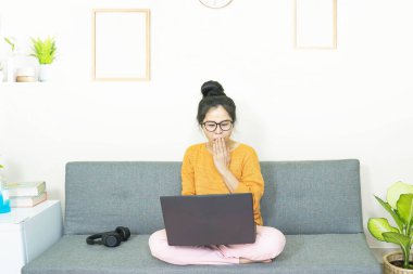 İnsanlar ve eğlence konsepti. Genç Asyalı güzel bir kadın gözlük takıyor, oturma odasında gri koltukta oturuyor, online yaşam tarzını karantina altında yaşıyor..