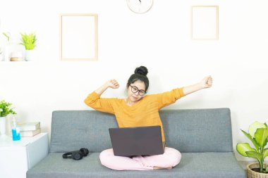 İnsanlar ve eğlence konsepti. Genç Asyalı güzel bir kadın gözlük takıyor, oturma odasında gri koltukta oturuyor, online yaşam tarzında çalışıyor, evde karantina altında kahveyle rahatlıyor.