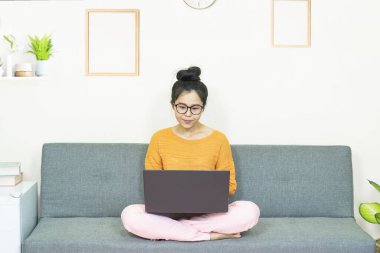 İnsanlar ve eğlence konsepti. Genç Asyalı güzel bir kadın gözlük takıyor, oturma odasında gri koltukta oturuyor, online yaşam tarzında çalışıyor, karantina altında evinde dinleniyor.