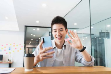 Ofiste sıradan gömlekli Asyalı genç iş adamı, video konferansında iş arkadaşlarını selamlıyor..