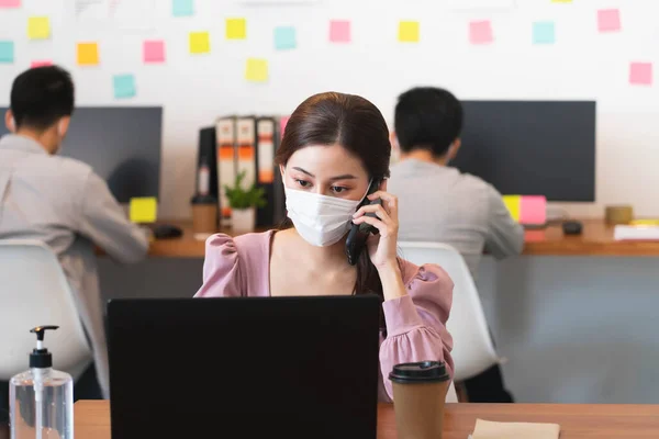 Seçici odak noktası, koruyucu yüz maskesi takan Asyalı güzel iş kadını. Ofiste cep telefonuyla konuşuyor. Covid-19 enfeksiyonunu önlemek için normal yaşam tarzıyla çalışıyor..