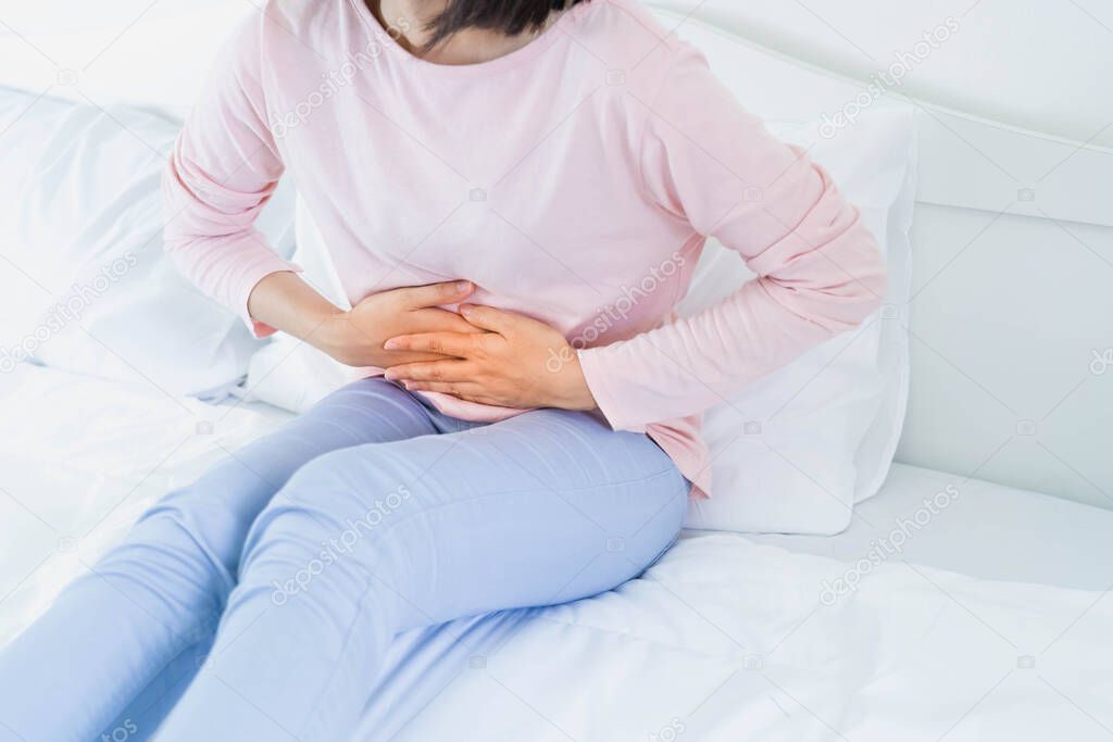 View of young woman suffering from stomachache on bed at home holding hands on her stomach having painful  stomach ache in bedroom, Menstrual period or abdominal pain