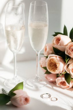 Alyanslar iki kadeh şampanya ve güneşte bir buket gülün yanında.