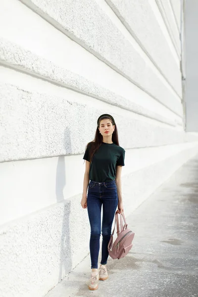 Brunette girl in jeans, a green T-shirt, a hair band and a pink backpack stands against a white wall