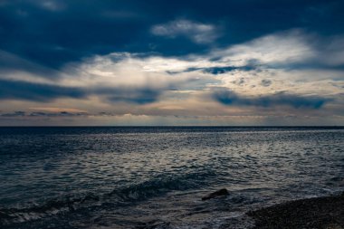 Deniz üzerinde bulutlu bir akşam gökyüzü, güneş ışığı bulutların ve sislerin arasından kırılıyor, bir çakıl taşı kıyısında dalgalar akıyor.