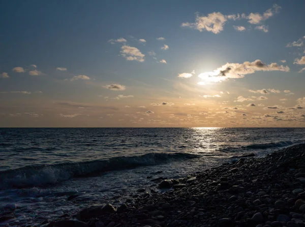 Sunset seascape, sunlight track along the water, sun in the clouds, wave runs on the pebble shore