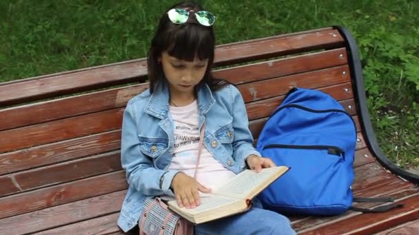 Κορίτσι μαθήτρια με σακίδιο κάθεται σε ένα παγκάκι και διαβάζει ένα βιβλίο — Αρχείο Βίντεο