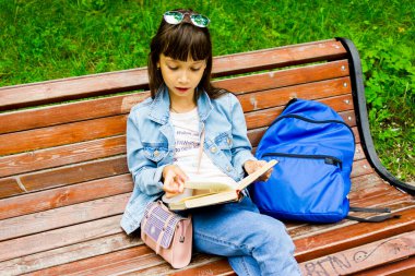 Güzel ve mutlu genç öğrenci kız bankta oturuyor, elleri kitap yığınının üzerinde, kameraya bakıyor ve gülümsüyor. Arka planda yaz veya bahar yeşil parkı