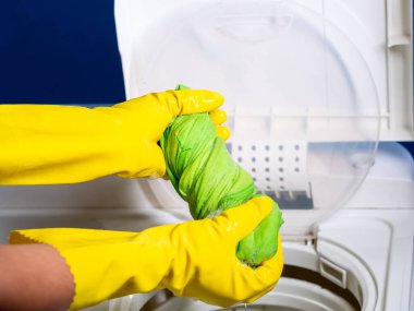 Sarı eldivenli, yeşil gömleği yarı otomatik çamaşır makinesinden köpükle sıkan bir kızın elleri.