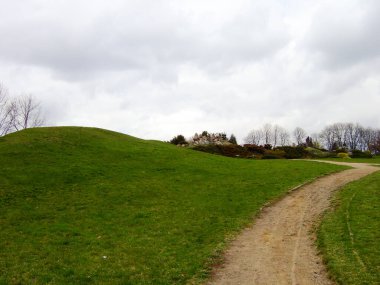 On the left is a green grassy hill and near it a dirt track against a gray sky with clouds. In the background are shit