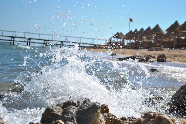 Las olas del mar golpean contra la orilla con spray. En el fondo hay una playa, un muelle que prohíbe nadar una bandera roja — Foto de Stock