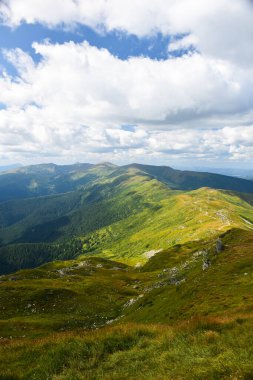 Yeşil çimlerle kaplı sırt, yukarıdan çekilen güneş tarafından parlak bir şekilde aydınlatılır. Üzerinde bulutlu mavi bir gökyüzü var..