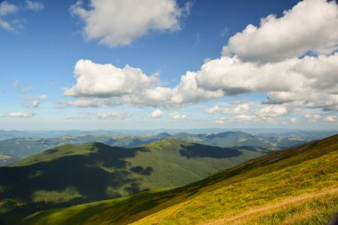 Yeşil çimlerle kaplı sırt, yukarıdan çekilen güneş tarafından parlak bir şekilde aydınlatılır. Üzerinde bulutlu mavi bir gökyüzü var..