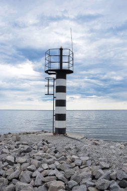 Deniz kıyısında küçük demir bir deniz feneri, taşlarla dolu, bulutlu mavi bir gökyüzüne karşı