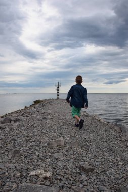 Sırtı dönük bir çocuk taştan yapılmış bir iskele boyunca uzak bir deniz fenerine doğru yürüyordu. Gökyüzü bulutlarla kaplı.