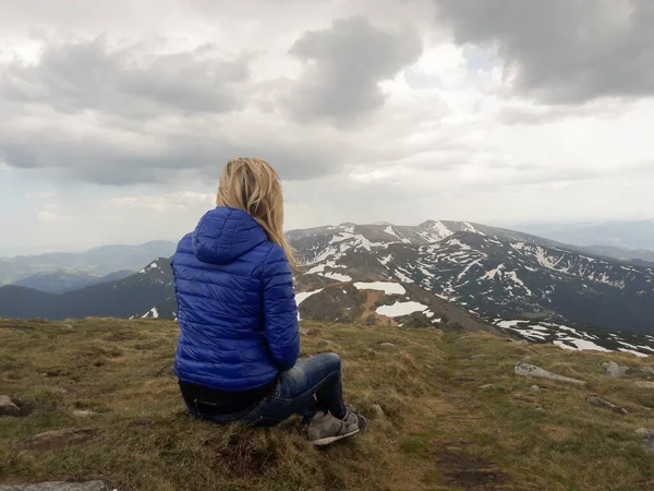 Ceketli sarışın bir kız sırtı yüksek bir dağda oturuyor. Bulutların ve yüksek dağların ardında