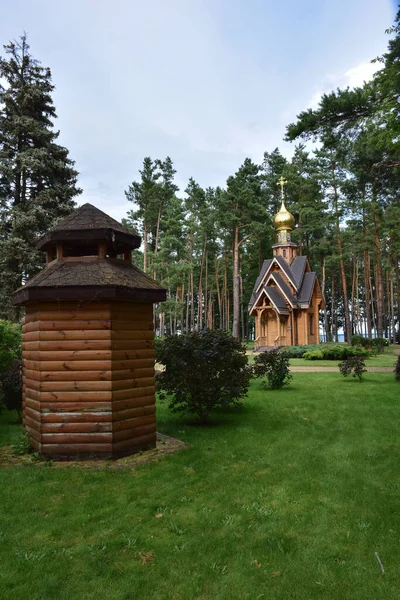 V borovém letním lesoparku se nachází malý dřevěný křesťanský kostelík. V popředí je dřevěný dům bez oken. — Stock fotografie
