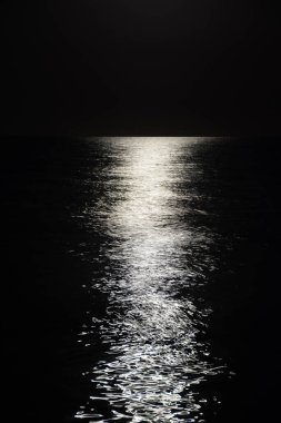Ayın parlak yansıması karanlık bir gecede sakin deniz dalgaları üzerinde ufukta beliriyor. Siyah ve beyaz.