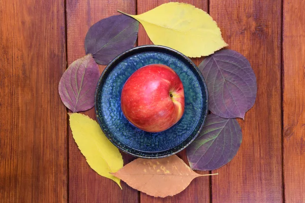 На коричневом деревянном фоне лежит тарелка с красным яблоком на ней. Осень красочные листья лежат вокруг блюдца в кругу. — стоковое фото