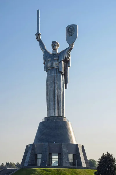 A huge iron statue of the Motherland Mother stands on a hill. In one hand, a sword raised up, in the other, a shield. Photographed during the day against the blue sky.