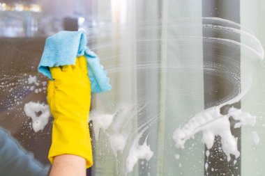 Sarı lastik eldivenli bir el odanın camını siler.