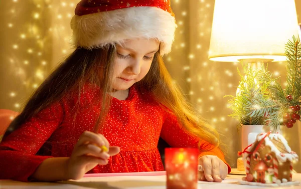 Kırmızı Noel şapkalı, kırmızı elbiseli küçük bir kızın portresi. Noel Baba 'ya mektup yazıyor. Bir Noel çelenginin arkasında.