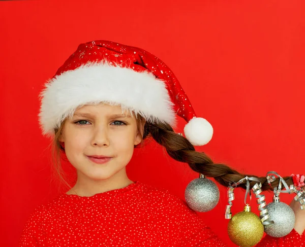 Mutlu Noeller ve mutlu yıllar. Kırmızı elbiseli ve Noel Baba şapkalı güzel bir kızın portresi. Üzerinde Noel topları asılı at kuyruğu var. kırmızı arka plan. Metin için boşluk.
