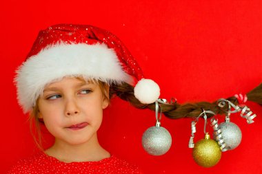 Mutlu Noeller ve mutlu yıllar. Kırmızı elbiseli ve Noel Baba şapkalı güzel bir kızın portresi. Üzerinde Noel topları asılı at kuyruğu var. kırmızı arka plan. Metin için boşluk.