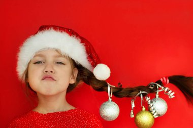 Mutlu Noeller ve mutlu yıllar. Kırmızı elbiseli ve Noel Baba şapkalı güzel bir kızın portresi. Üzerinde Noel topları asılı at kuyruğu var. kırmızı arka plan. Metin için boşluk.