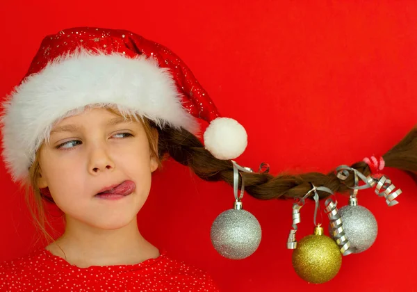 Mutlu Noeller ve mutlu yıllar. Kırmızı elbiseli ve Noel Baba şapkalı güzel bir kızın portresi. Üzerinde Noel topları asılı at kuyruğu var. kırmızı arka plan. Metin için boşluk.