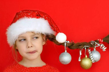 Mutlu Noeller ve mutlu yıllar. Kırmızı elbiseli ve Noel Baba şapkalı güzel bir kızın portresi. Üzerinde Noel topları asılı at kuyruğu var. kırmızı arka plan. Metin için boşluk.