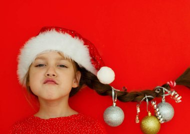 Mutlu Noeller ve mutlu yıllar. Kırmızı elbiseli ve Noel Baba şapkalı güzel bir kızın portresi. Üzerinde Noel topları asılı at kuyruğu var. kırmızı arka plan. Metin için boşluk.