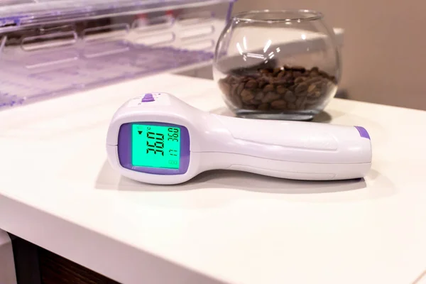 Non-contact infrared thermometer lies on the table and shows normal temperature 36.0. Nearby lies a glass bowl with coffee beans. Safety at work theme.
