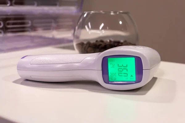 Non-contact infrared thermometer lies on the table and shows normal temperature 36.0. Nearby lies a glass bowl with coffee beans. Safety at work theme.