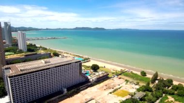 Tayland 'ın deniz kıyısındaki prestijli beyaz apartmanlardan havadan 4K manzara. Yakınlarda tekneli bir liman var. Kıyıda yüzme havuzları ve büyüyen palmiye ağaçları var. Ufukta yüksek dağlar görülebilir. Panorama