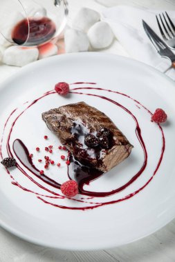 Beyaz tabakta ahududu soslu geyik eti. Beyaz ahşap bir masada.