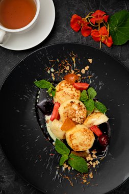 Karanlık bir arka planda siyah tabakta soslu, böğürtlenli ve naneli Ukrayna peynirli kekleri.