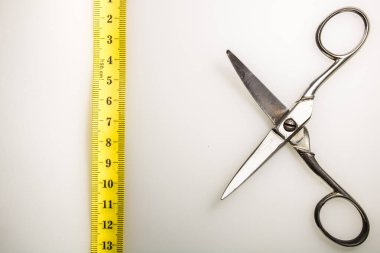 Metal makas, açık bir pozisyonda, sarı şeritli bir ölçünün önünde, beyaz bir arkaplanda konumlandırılmış.