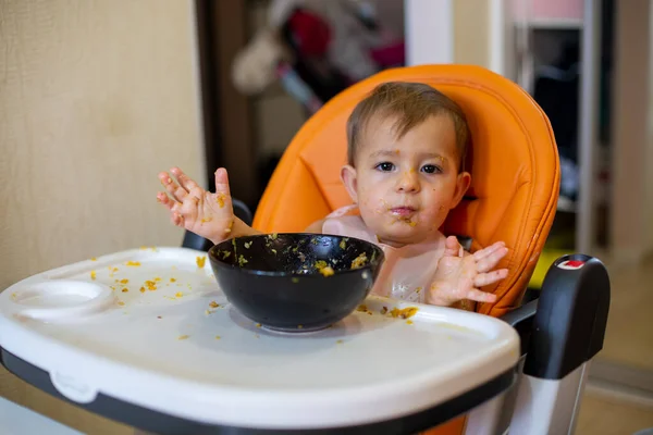 Linda niña de un año en un asiento de niño naranja delante de un plato de comida arroja sus manos y mira a la cámara. todo alrededor en la comida. enfoque suave, primer plano, fondo borroso — Foto de Stock