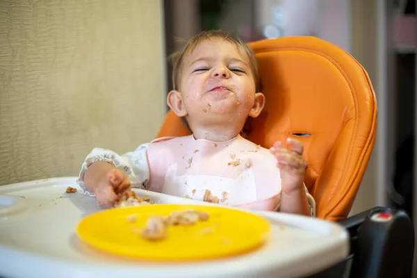 Problema alimentar al bebé. Lindo bebé en un asiento de niño naranja mira a la cámara y sonríe. migas y un plato naranja sobre la mesa. primer plano, vista frontal, enfoque suave, fondo borroso — Foto de Stock