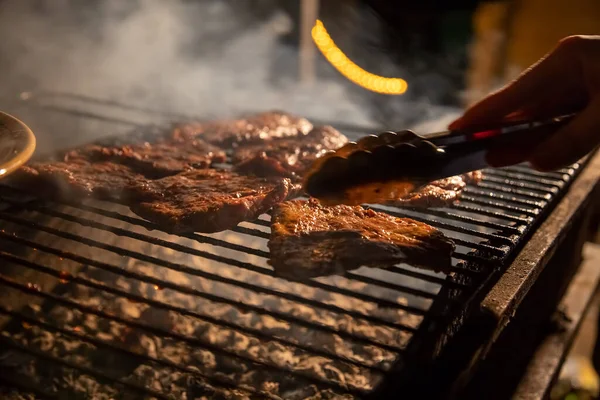 Večer gril na grilu, na kterém se grilují chutné šťavnaté steaky nad otevřeným ohněm, ruka drží kleště a převrací maso. zblízka, měkké soustředění. kouř je zvýrazněn — Stock fotografie
